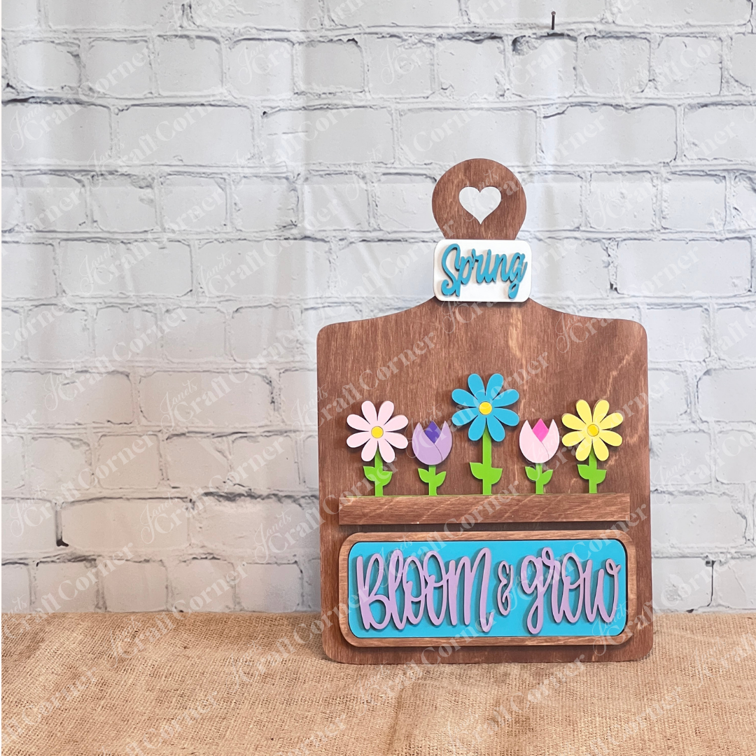 The Bloom & Grow Interchangeable Insert kit from Janet's Craft Corner, featuring colorful flowers and "Bloom & Grow" in blue on brown, complemented by a charming "Spring Blooms" insert against a white brick wall, adds a decorative touch with its 3-piece DIY home decor craft set.