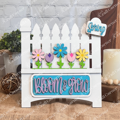 Janet's Craft Corner presents the "Bloom & Grow Interchangeable Insert" kit, featuring decorative signs with a white picket fence, colorful flowers, and "Bloom & Grow" in blue and pink. Includes a tag reading "Spring Blooms," ideal for adding charm to any setting alongside bricks, lush plants, a woven ball, or candle.