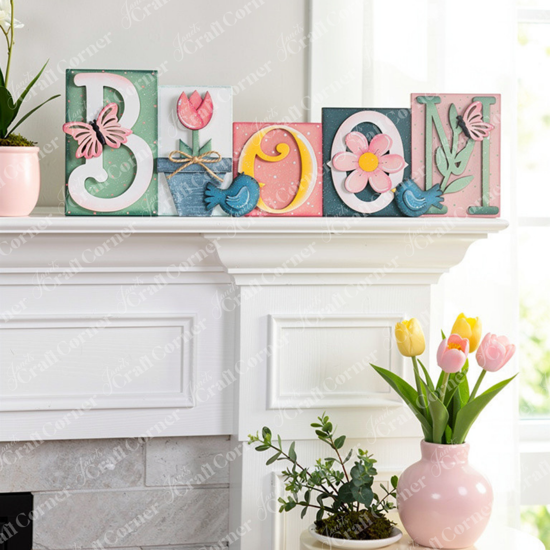 The Bloom Word Block by Janet's Craft Corner spells "BLOOM" with charming floral and butterfly motifs, including a tulip, daisy, and pastel-colored butterflies. This piece,shown displayed on a mantle, is perfect for enhancing home décor.