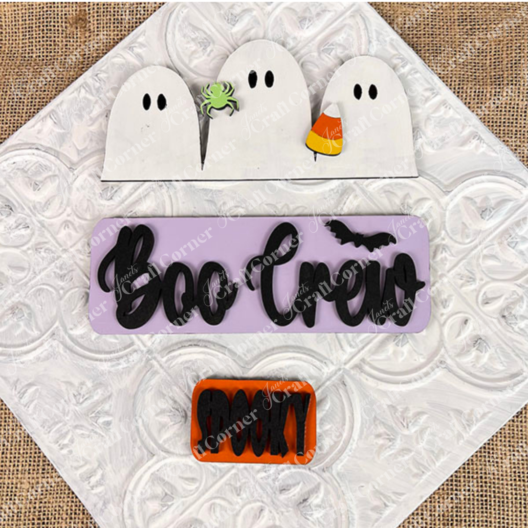 The Boo Crew Interchangeable Insert DIY home decor kit from Janet's Craft Corner features Halloween-themed decorations on a textured background, including three white ghosts, a green spider with candy corn, and a purple plaque with "Boo Crew" in black text accompanied by a bat. Completing the set is an orange plaque reading "Spooky.