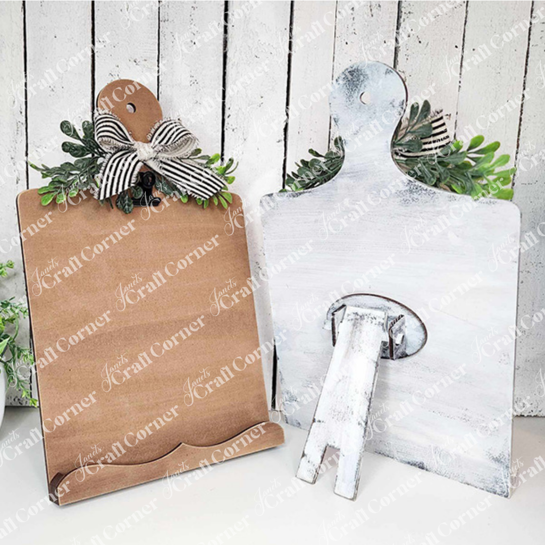 Two Breadboard Sign Holders from Janet's Craft Corner, one in a plain brown finish and the other whitewashed, each featuring leafy embellishments and a striped bow at the top, are displayed against a rustic white wooden backdrop. Perfect for farmhouse decor enthusiasts or as part of your DIY home decor craft kit.