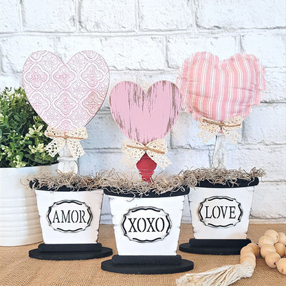 Introducing the Bucket Heart Trio from Janet's Craft Corner: a delightful DIY kit featuring three decorative pots, each topped with a heart labeled "AMOR," "XOXO," and "LOVE." These hearts showcase various pink patterns and textures. They are ideal for charming home décor, displayed against a white brick background with greenery on the left.