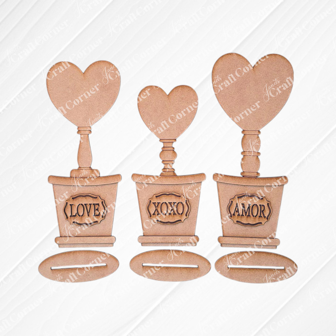 Introducing the Bucket Heart Trio from Janet's Craft Corner: a delightful DIY kit featuring three decorative pots, each topped with a heart labeled "AMOR," "XOXO," and "LOVE." They are ideal for charming home décor, displayed against a white wood background.