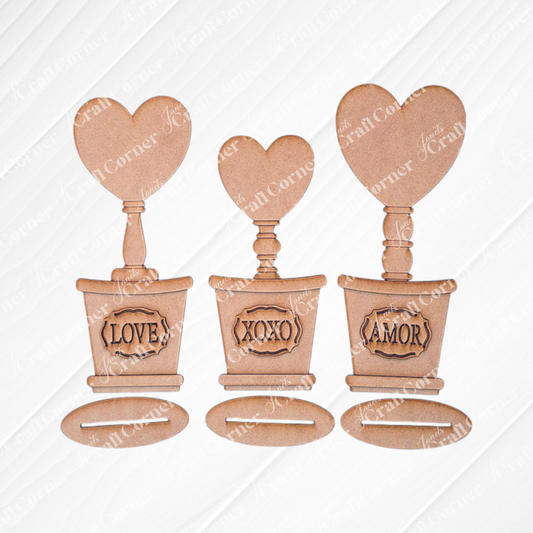 Introducing the Bucket Heart Trio from Janet's Craft Corner: a delightful DIY kit featuring three decorative pots, each topped with a heart labeled "AMOR," "XOXO," and "LOVE." They are ideal for charming home décor, displayed against a white wood background.