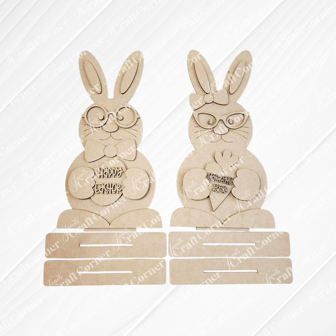 Janet's Craft Corner offers a DIY seasonal home decor kit featuring the Bunny Couple Shelf Sitters. These charming wooden Easter bunnies include one with glasses and a bowtie holding a "Happy Easter" egg, and another with glasses holding a carrot-shaped "Feliz Páscoa" piece against a slatted background.