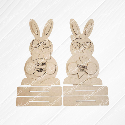 Janet's Craft Corner offers a DIY seasonal home decor kit featuring the Bunny Couple Shelf Sitters. These charming wooden Easter bunnies include one with glasses and a bowtie holding a "Happy Easter" egg, and another with glasses holding a carrot-shaped "Feliz Páscoa" piece against a slatted background.
