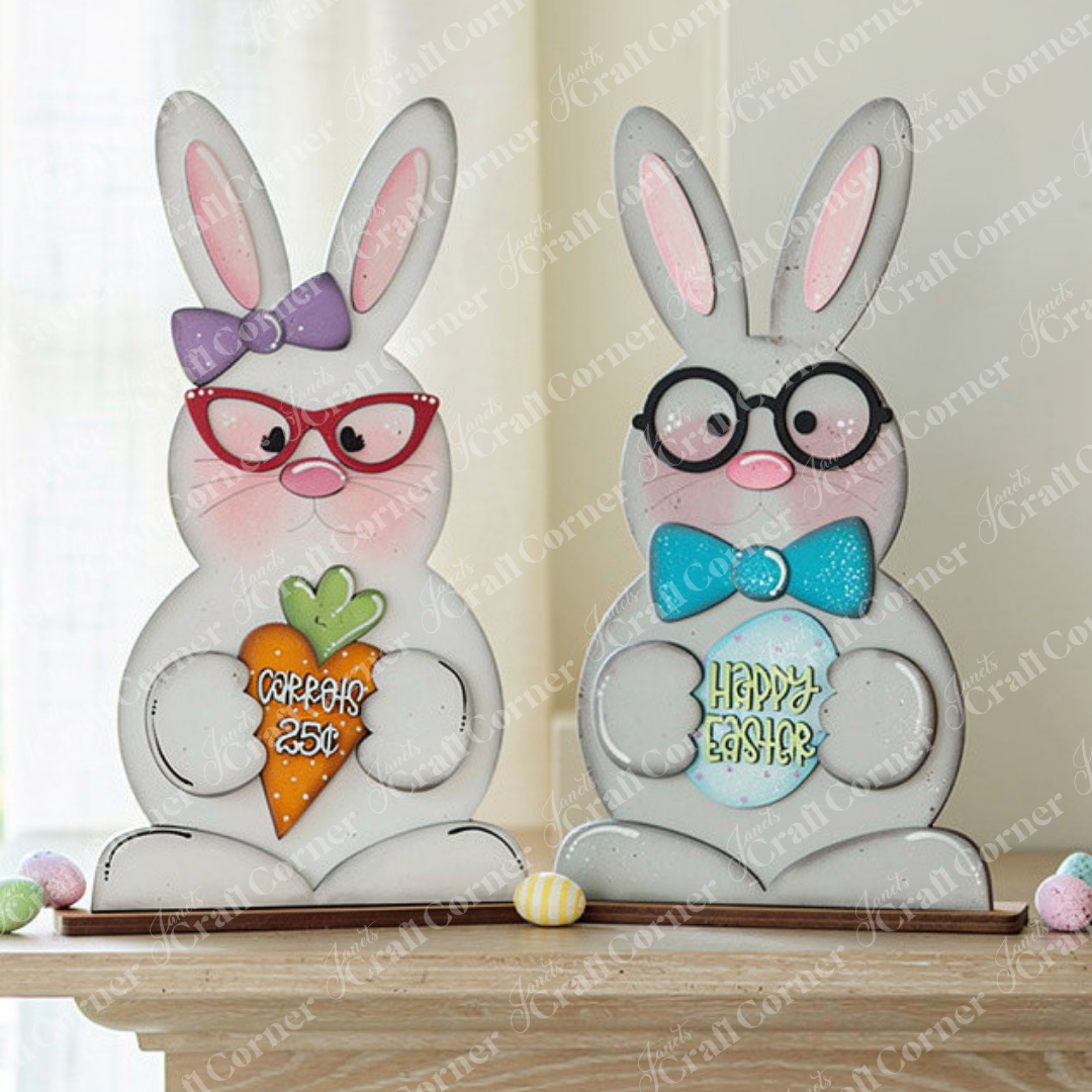 Janet's Craft Corner presents the delightful Bunny Couple Shelf Sitters, which feature two charming figures wearing glasses. One bunny holds a carrot sign inscribed with "SPRING 25¢," and the other displays an egg sign that says "HAPPY EASTER." Their adorable expressions and pastel accents provide a lovely contrast against a light background, adding cheer to your home decor.