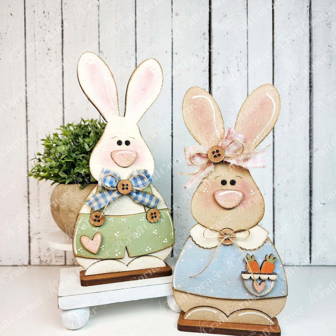 Janet's Craft Corner Bunny Couple Shelf Sitters DIY kit includes two wooden bunny figures, each with painted faces and pastel outfits—one in green with a blue bow, the other in blue with a pink bow and carrot tray—perfect for spring decorating.