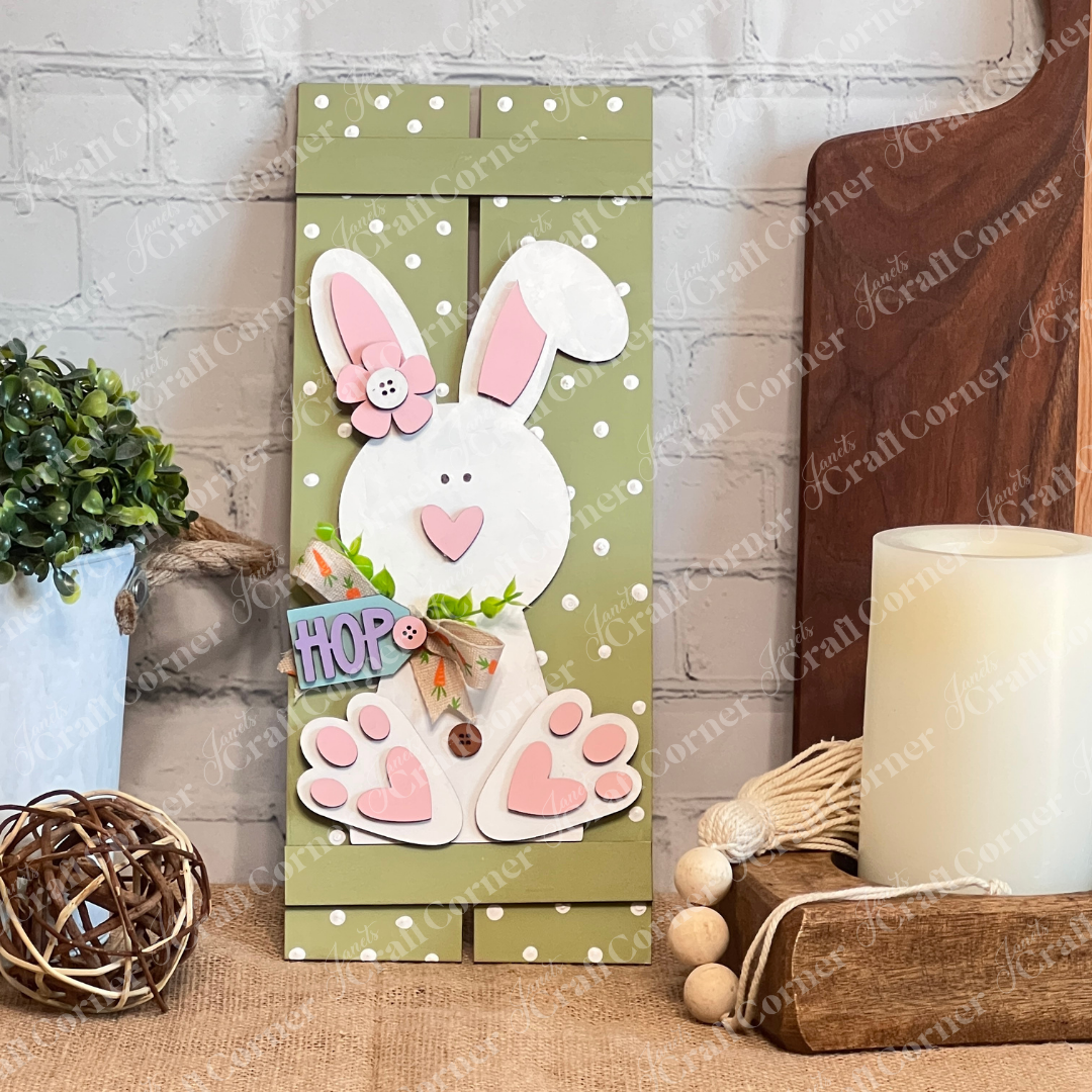 Janet's Craft Corner's Bunny Table Top Pallet craft kit features a wooden Easter bunny with a pink nose and button eyes, holding a "HOP" sign beside a green potted plant, a breadboard, and a candle. Perfect as a shelf leaner or mantle décor piece, it adds charm with its light-colored background.