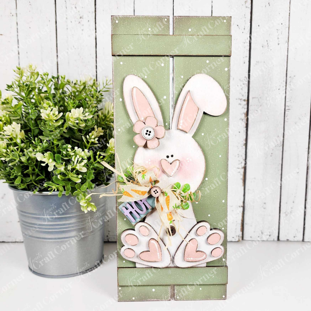 Janet's Craft Corner's Bunny Table Top Pallet craft kit features a wooden Easter bunny with a pink nose and button eyes, holding a "HOP" sign beside a green potted plant. Perfect as a shelf leaner or mantle décor piece, it adds charm with its light-colored background.