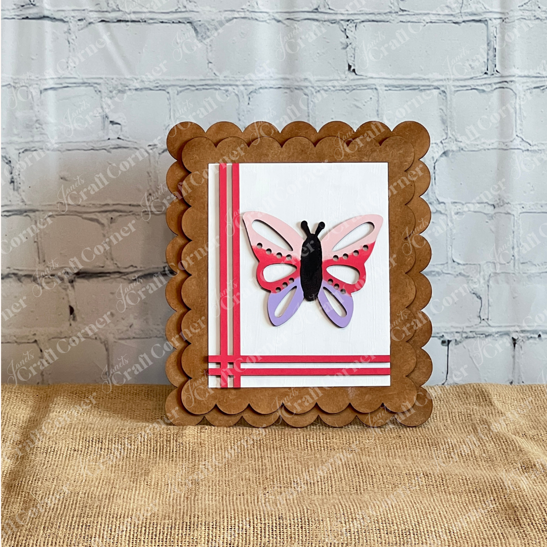 A square, scalloped-edged brown frame from Janet's Craft Corner’s Spring Switchable Farmhouse Signs DIY Kit holds a striped white card and layered butterfly—ideal for seasonal farmhouse décor or tiered tray styling against a white brick wall.