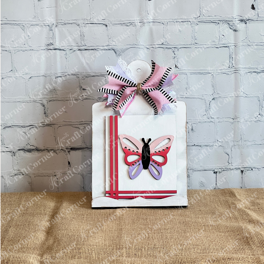 A white clipboard with a pink and black butterfly, diagonal pink stripes, and a layered bow from Janet's Craft Corner pairs perfectly with the Spring Switchable Farmhouse Signs DIY Kit for stylish seasonal decor against a white brick wall.