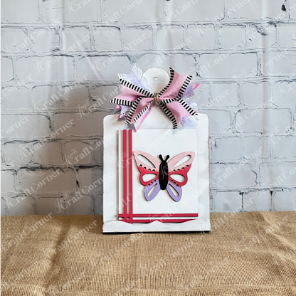A white clipboard with a pink and black butterfly, diagonal pink stripes, and a layered bow from Janet's Craft Corner pairs perfectly with the Spring Switchable Farmhouse Signs DIY Kit for stylish seasonal decor against a white brick wall.