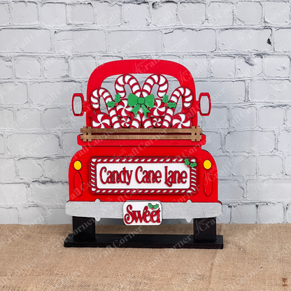 Displayed against a white brick wall is a decorative red truck adorned with candy canes and green bows, showcasing Janet's Craft Corner's hand-painted DIY home decor kit, the "Candy Cane Lane Interchangeable Insert," featuring signs that read "Candy Cane Lane" and "Sweet.