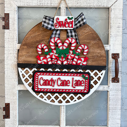 This "Candy Cane Lane" interchangeable insert from Janet's Craft Corner is a vibrant hand-painted element in a DIY home decor kit. It showcases vivid red and white candy cane decorations, complete with a black and white checkered bow. The word "Sweet" is beautifully highlighted at the top, surrounded by holly accents against a rustic wooden door backdrop.