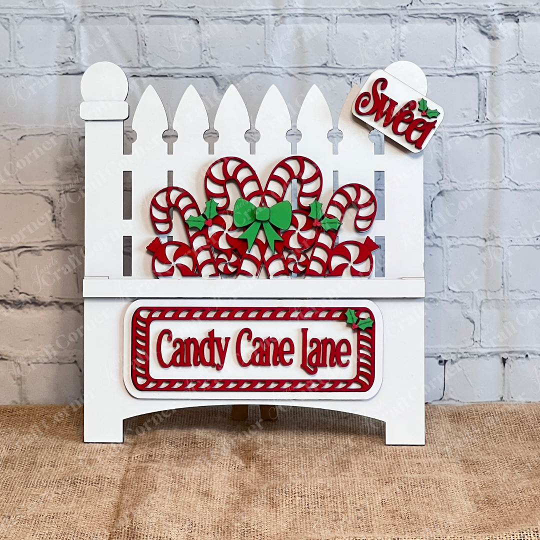 Displayed against a white brick wall is a decorative white garden fence adorned with candy canes and green bows, showcasing Janet's Craft Corner's hand-painted DIY home decor kit, the "Candy Cane Lane Interchangeable Insert," featuring signs that read "Candy Cane Lane" and "Sweet.
