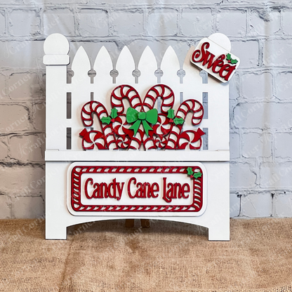 Displayed against a white brick wall is a decorative white garden fence adorned with candy canes and green bows, showcasing Janet's Craft Corner's hand-painted DIY home decor kit, the "Candy Cane Lane Interchangeable Insert," featuring signs that read "Candy Cane Lane" and "Sweet.
