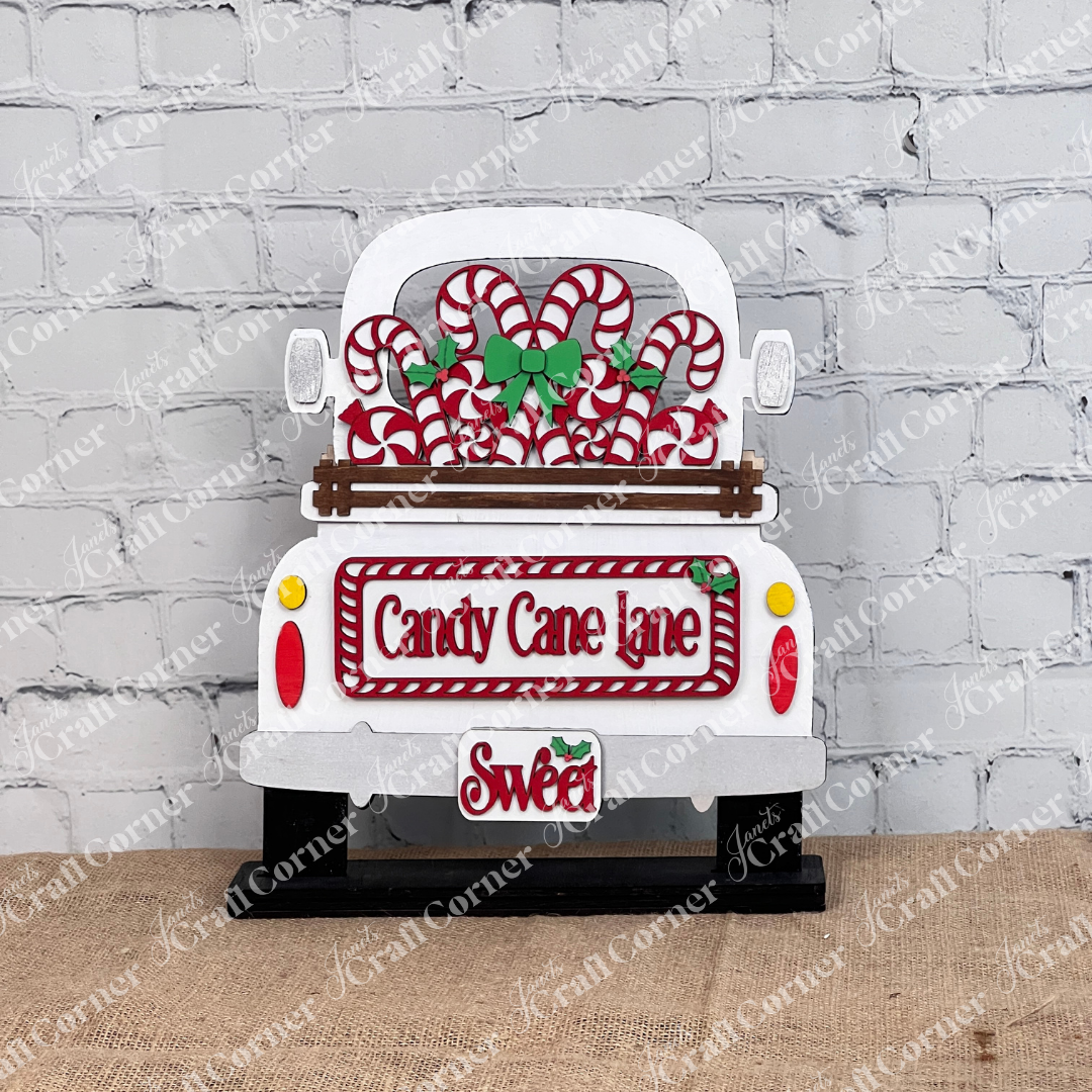 Displayed against a white brick wall is a decorative white truck adorned with candy canes and green bows, showcasing Janet's Craft Corner's hand-painted DIY home decor kit, the "Candy Cane Lane Interchangeable Insert," featuring signs that read "Candy Cane Lane" and "Sweet.
