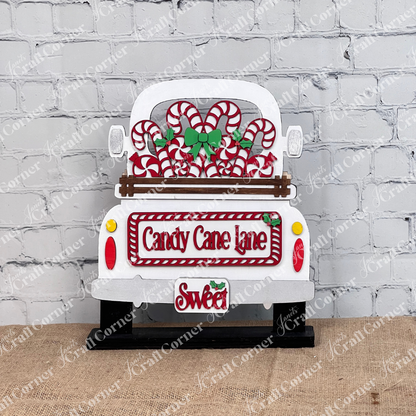 Displayed against a white brick wall is a decorative white truck adorned with candy canes and green bows, showcasing Janet's Craft Corner's hand-painted DIY home decor kit, the "Candy Cane Lane Interchangeable Insert," featuring signs that read "Candy Cane Lane" and "Sweet.