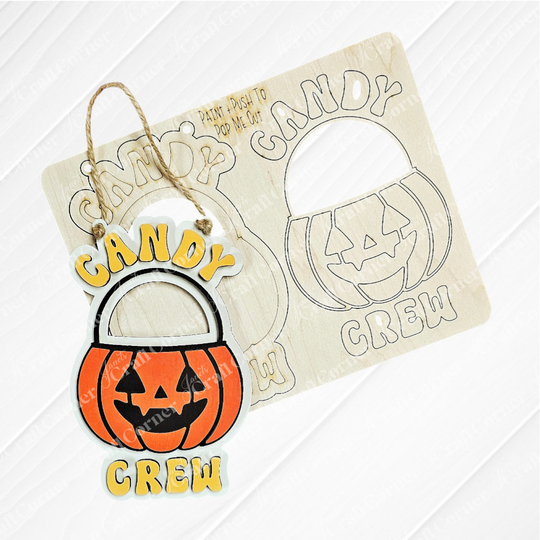 The Candy Crew Kids Pop-Out by Janet's Craft Corner is a wooden tree ornament shaped like a jack-o'-lantern bucket, shown with its cutout template. With orange, black, yellow, and white colors, it's perfect for festive holiday decor.