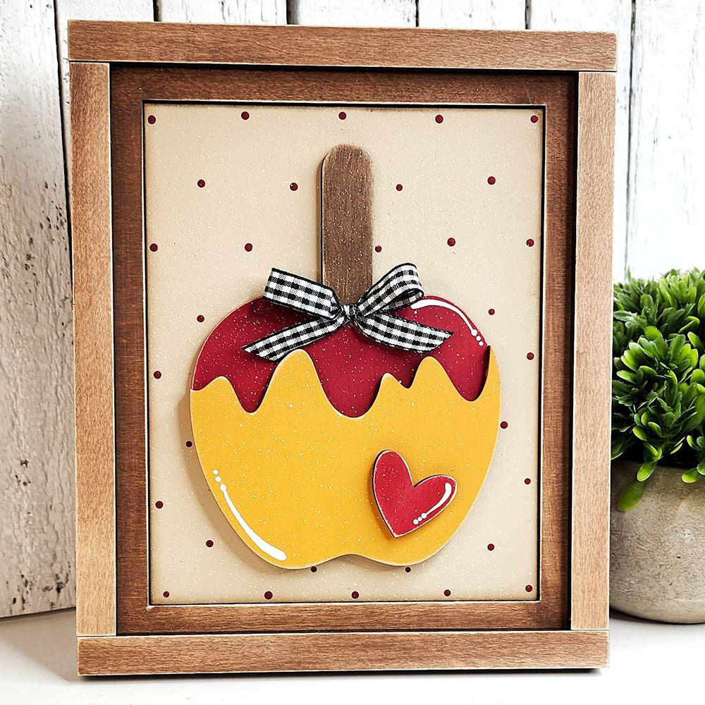 This Caramel Apple Interchangeable Sign, part of the DIY home decor craft kit by Janet's Craft Corner, features a framed design of a yellow apple with a red-dipped top and heart detail. It is elegantly adorned with a black and white checkered bow on the wooden stem. Displayed against a dotted backdrop, this sign stands on a wooden base accompanied by a small plant, making it ideal for interchangeable home décor.