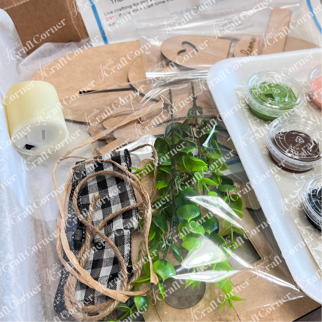 A close-up of Janet's Craft Corner's Crafted by You! Subscription—monthly DIY home decor kit for February 2025 with faux greenery, paints, checked fabric, twine, wood shapes, and a tealight candle—all neatly packaged for craft lovers.