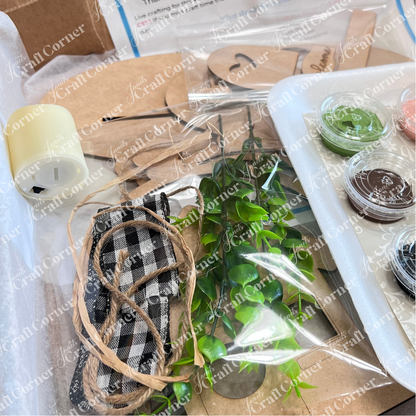 A close-up of Janet's Craft Corner's Crafted by You! Subscription—monthly DIY home decor kit for February 2025 with faux greenery, paints, checked fabric, twine, wood shapes, and a tealight candle—all neatly packaged for craft lovers.