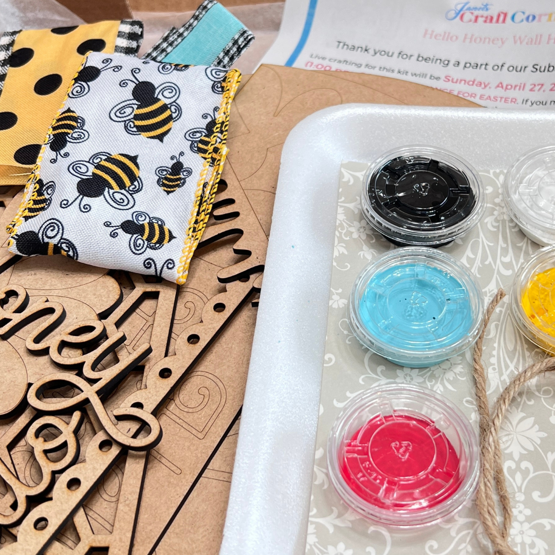 A close-up of Janet's Craft Corner Crafted by You! Subscription—DIY home decor kit—with "Honey" wooden cutouts, bee-patterned fabric, paint pots (black, yellow, blue, red), a paintbrush, and instructions in the background for the February 2025 Hello Honey Door Hanger kit.