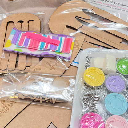 A Crafted by You! Subscription DIY home decor craft kit from Janet’s Craft Corner sits on the table, filled with wooden cutouts, colorful ribbon, and paint—perfect inspiration for your creative community.