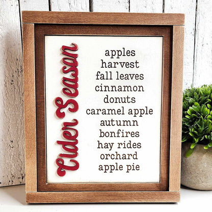 On the left, Janet's Craft Corner's Cider Words Interchangeable Sign, part of a DIY home decor craft kit, showcases "Cider Season" in vibrant red script. The right side displays cozy words such as "apples," "harvest," "fall leaves," and "cinnamon." A nearby small potted plant introduces a touch of nature to this charming craft insert scene.