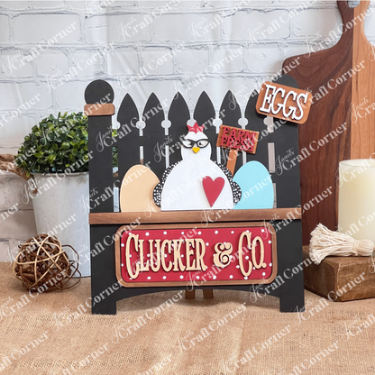 Janet's Craft Corner introduces the Clucker & Co. Interchangeable Insert - Painted, a decorative piece featuring a delightful chicken motif adorned with glasses, alongside a red heart and colorful eggs. The sign displays "EGGS" and "Farm Fresh" at the top, with "Clucker & Co." beneath on a black garden fence, making it an ideal choice for farmhouse decor enthusiasts seeking versatile craft inserts.
