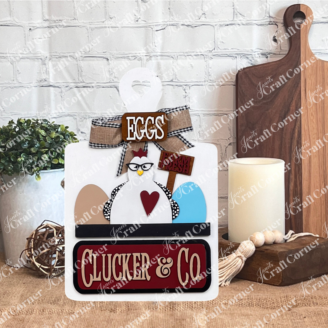 Janet's Craft Corner introduces the Clucker & Co. Interchangeable Insert - Painted, a decorative piece featuring a delightful chicken motif adorned with glasses, alongside a red heart and colorful eggs. The sign displays "EGGS" and "Farm Fresh" at the top, with "Clucker & Co." beneath on a white breadboard. making it an ideal choice for farmhouse decor enthusiasts seeking versatile craft inserts.