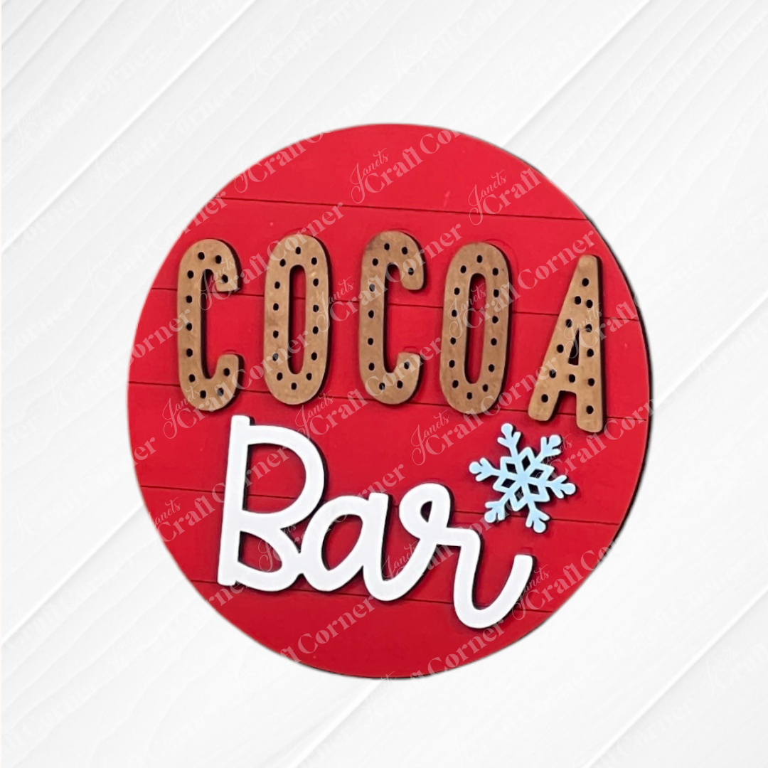 Janet’s Craft Corner’s Hot Cocoa Mini Shelf Sitters DIY kit features a round red sign with “COCOA” in brown uppercase, “Bar” in white script with a blue snowflake for the “a”—a perfect cozy seasonal decor accent.
