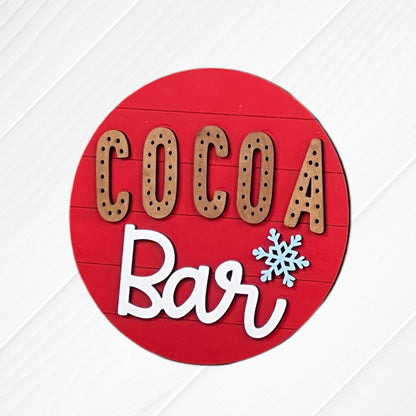 The Cup of Cheer Hot Cocoa Mini Shelf Sitters by Janet's Craft Corner features a round red sign with "COCOA Bar" in brown and white letters and a blue snowflake, perfect for hot cocoa decor on any white wooden background.
