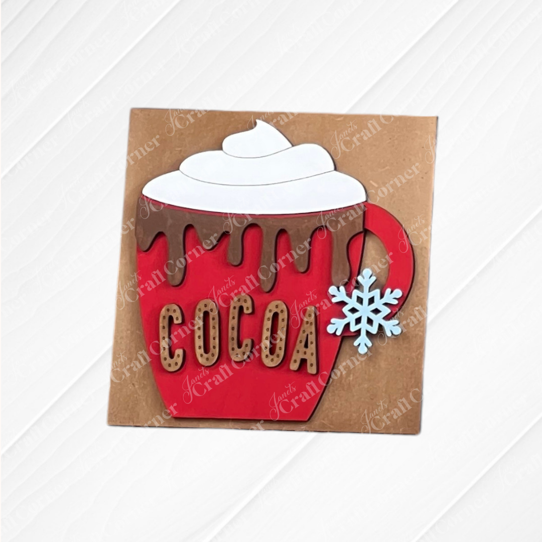 The Hot Cocoa Mini Shelf Sitter by Janet's Craft Corner is a red mug with chocolate drip, whipped cream top, and blue snowflake handle. "COCOA" decorates the front. Great for cozy DIY home decor or gifting. Set of 1 craft kit on a brown base.