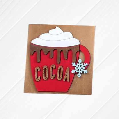 The Cup of Cheer Hot Cocoa Mini Shelf Sitter by Janet's Craft Corner features a red "COCOA" mug with whipped cream, chocolate drips, and a blue snowflake handle—perfect decor for your cocoa bar on a brown background.
