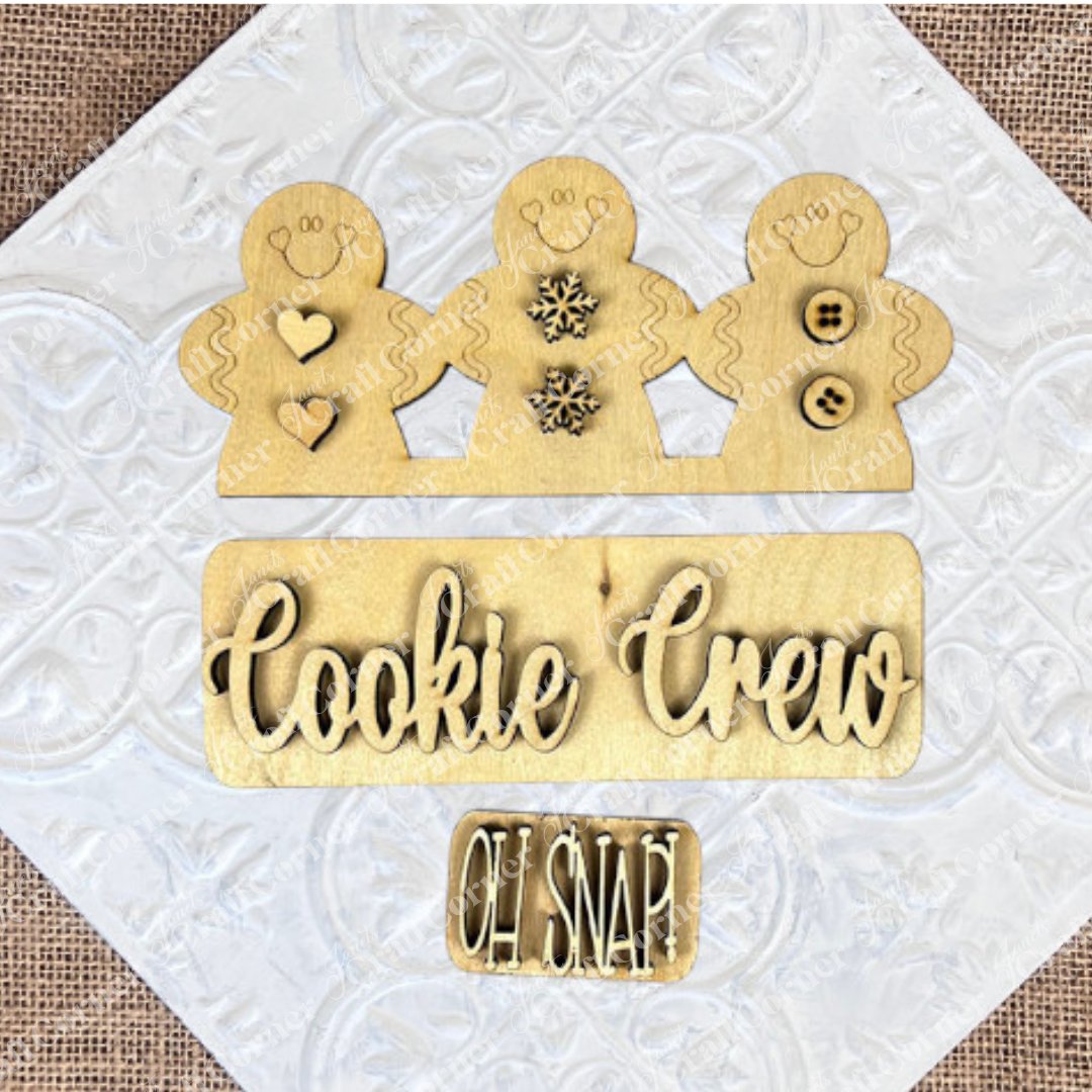 The Cookie Crew Interchangeable Insert by Janet's Craft Corner is an ideal DIY home decor kit, featuring wooden cutouts of three gingerbread men adorned with heart, snowflake, and button designs. A sign at the base displays "Cookie Crew" with a smaller "Oh Snap!" text, all crafted from light-colored wood.