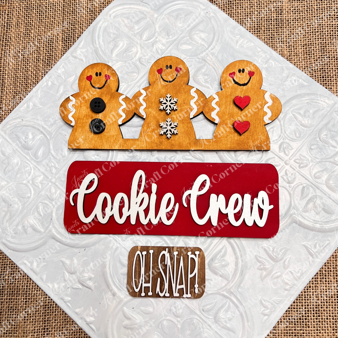 Three cheerful gingerbread figures—one with buttons, another with snowflakes, and the last with hearts—embellish a red sign that says "Cookie Crew," accompanied by a brown sign that reads "Oh Snap!" on a gray background. This charming DIY home decor kit from Janet's Craft Corner provides interchangeable decorations to brighten up the festive season.