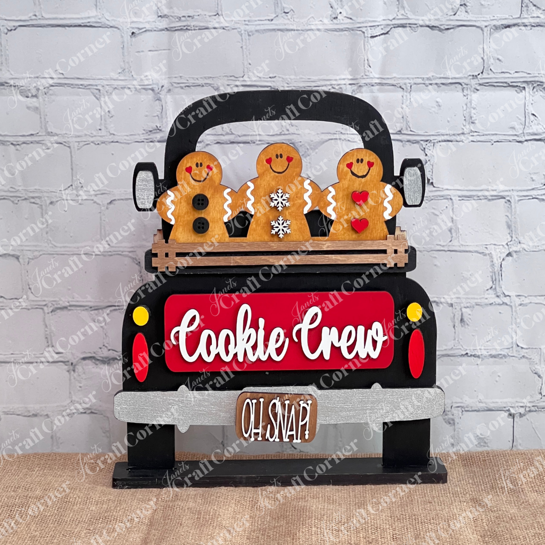 The Cookie Crew Interchangeable Insert from Janet's Craft Corner showcases three delightful gingerbread figures riding in a black truck labeled "Cookie Crew," with the whimsical phrase "Oh Snap!" underneath. Perfect as versatile decor, these gingerbread men are beautifully detailed with buttons, a snowflake, and hearts.