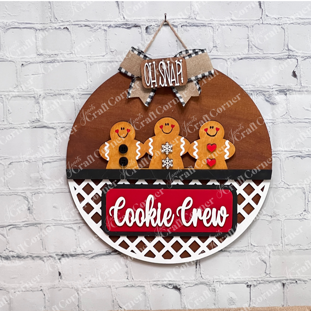 The Cookie Crew Interchangeable Insert by Janet's Craft Corner is a DIY seasonal decor kit with 3 pieces, featuring gingerbread men designs and festive "OH SNAP" and "Cookie Crew" text on a white brick wall background.