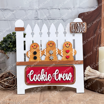 The Cookie Crew Interchangeable Insert by Janet's Craft Corner is a DIY home decor craft kit featuring three uniquely patterned gingerbread men on a white picket fence. The board reads "Oh Snap!" and includes a red "Cookie Crew" sign against a white brick wall, perfect for seasonal inserts.