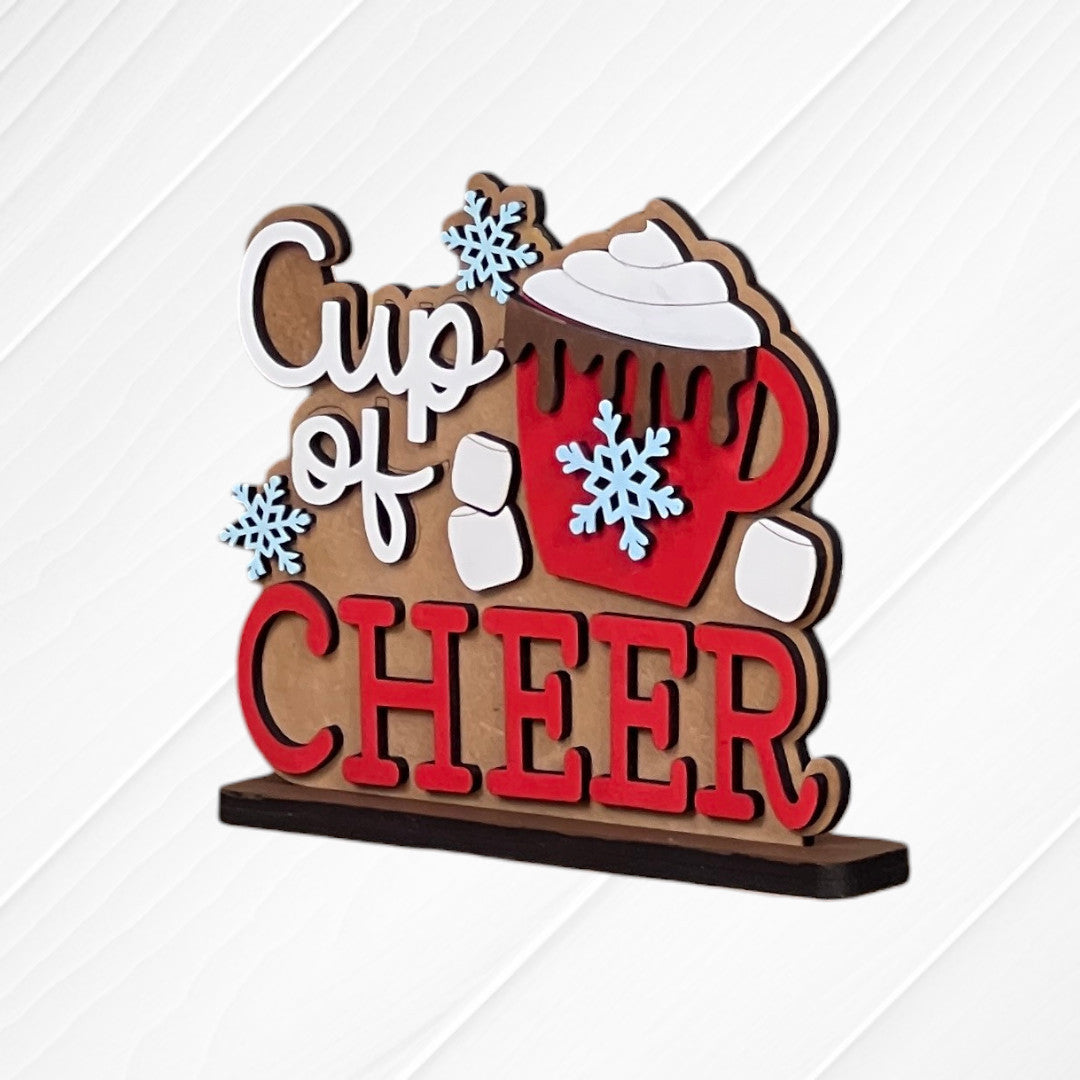 Janet's Craft Corner Cup of Cheer Hot Cocoa Mini Shelf Sitters feature a red mug with whipped cream, snowflakes, and marshmallows on a white wood background. "Cup of CHEER" is printed in bold letters—perfect festive decor for your cocoa bar.
