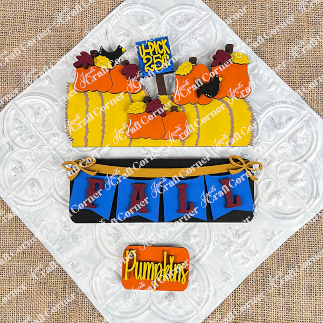 A set of beautifully decorated cookies displayed on a woven mat captures the essence of fall décor with designs featuring pumpkins, hay bales, and crows. One cookie displays the message "4-Pick 5 for 25¢" in a cozy autumn atmosphere, perfectly embodying the style of Janet's Craft Corner's Fall Hay Bale Interchangeable Insert - Painted. This scene brings an interchangeable home decor touch to your seasonal festivities.