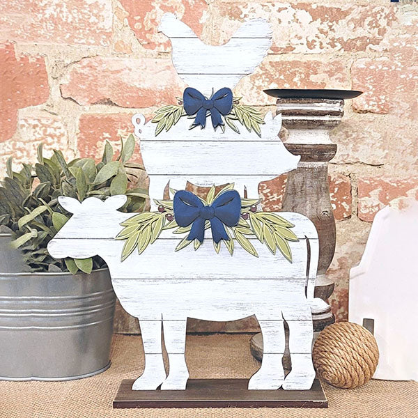 Introducing the Farm Animal Stacker Shelf Sitter from Janet's Craft Corner, a delightful home décor piece. This rustic wooden farmhouse decoration features gracefully stacked silhouettes of a cow, pig, and chicken. Each animal is adorned with blue bows and green leaf garlands, perfectly complemented by the brick wall backdrop and an accompanying potted plant for a complete pastoral charm.