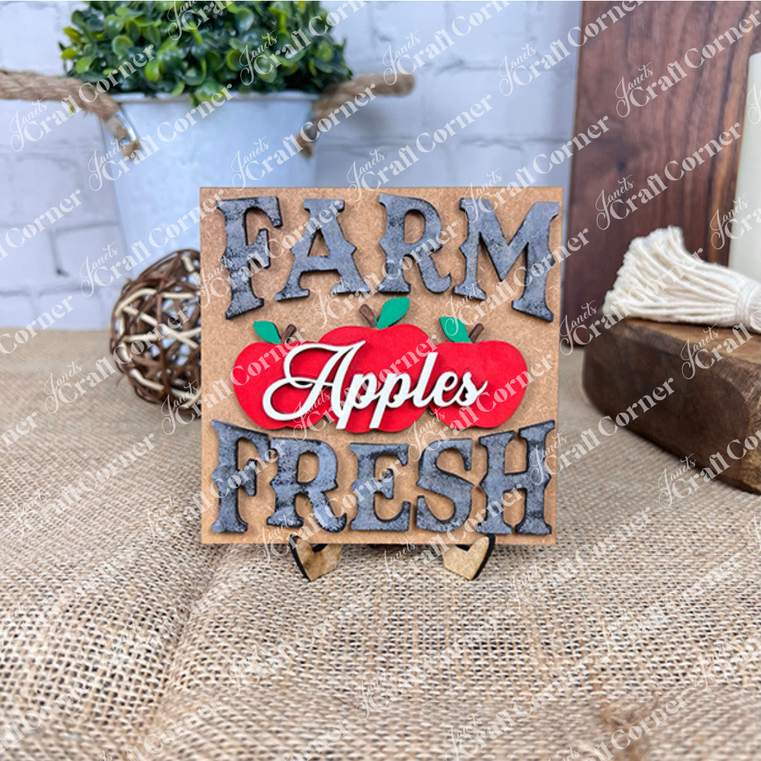 Janet's Craft Corner Apple Swappable Squares DIY kit features a "Farm Fresh Apples" decorative sign with a red apple illustration, wood cut-outs, rustic burlap, greenery, and interchangeable pieces for charming seasonal home decor.