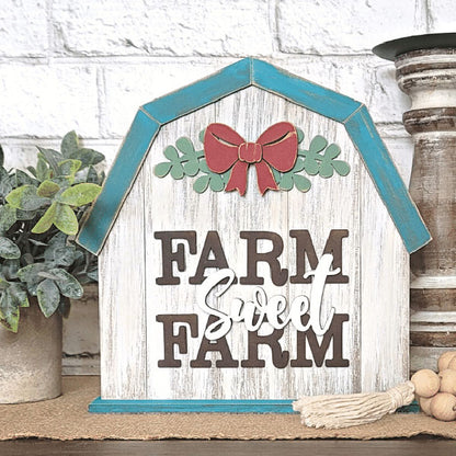 The "Farm Sweet Farm Shelf Sitter" from Janet's Craft Corner is a decorative sign in the shape of a barn with blue trim, adorned with a red bow and green leaves at the top. It features the words "Farm Sweet Farm," with "Sweet" elegantly scripted. This charming piece of home décor is beautifully displayed on a rustic table alongside potted plants and a wooden tassel.