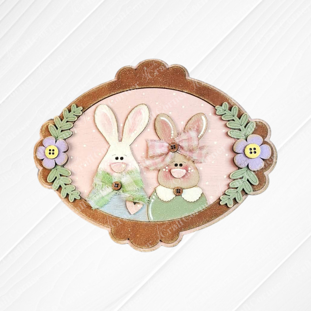 Janet’s Craft Corner Framed Bunny Couple Door/Wall Hanger—DIY home decor kit (set of 1) with button-nosed bunnies in green and pink, purple flowers, and green leaves on a light pink background.
