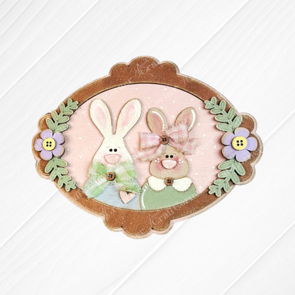 Janet’s Craft Corner Framed Bunny Couple Door/Wall Hanger—DIY home decor kit (set of 1) with button-nosed bunnies in green and pink, purple flowers, and green leaves on a light pink background.