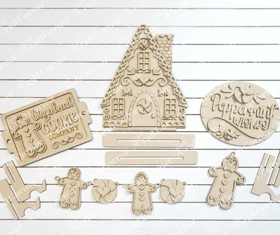 The Gingerbread Decor Set from Janet's Craft Corner is a 5-piece DIY home decor craft kit that includes charming hand-painted wooden gingerbread-themed pieces. The set features a decorative house, gingerbread people, candy shapes, and plaques with engraved designs, all beautifully displayed on a white wooden background.