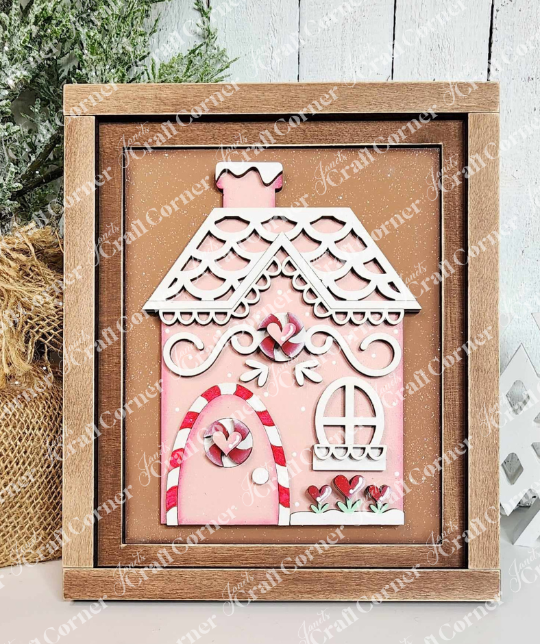 A painted sample of the Gingerbread House Switchable Farmhouse Sign and Farmhouse frame from Janet's Craft Corner's displayed against a white shiplap wall. The Gingerbread Switchable Farmhouse Sign features a cute house with red & white peppermint and white icing trim on a cinnamon background in a wooden frame—ideal for seasonal home decor. 